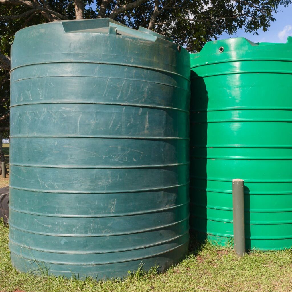 Two large, green water storage tanks stand side by side on a grassy patch under a clear sky