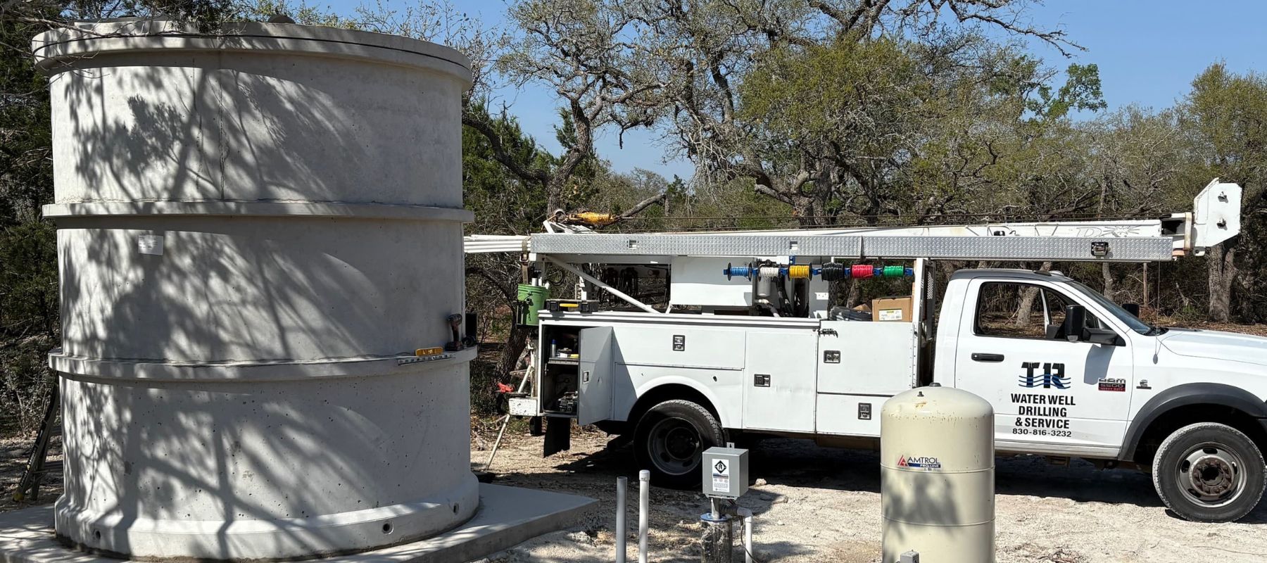 A tall, stacked concrete water tank stands beside a "TR WATER WELL DRILLING & SERVICE" truck and a beige water well system