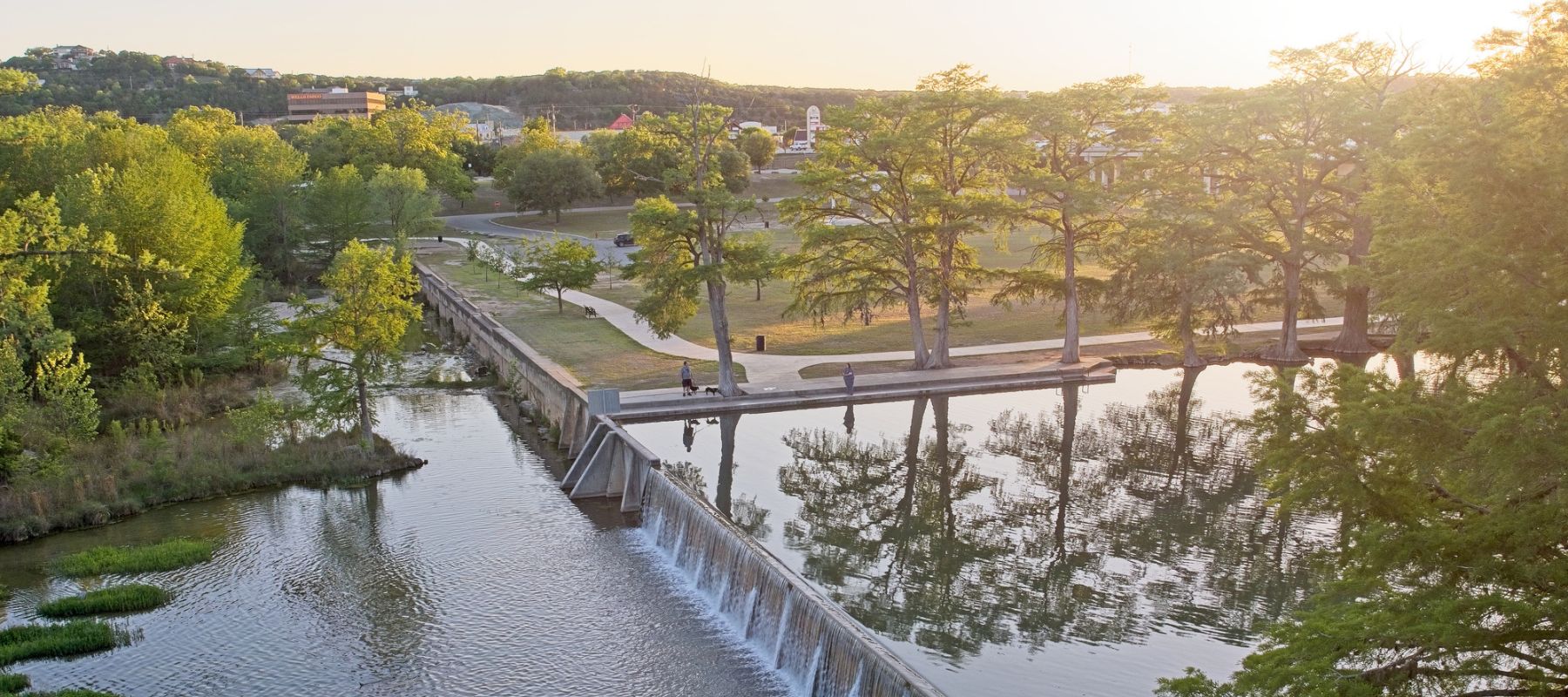 A riverside park in Kerrville, Texas, with a walkway, trees, and a small dam at sunset
